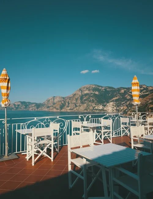 Terrazza con sedie e tavoli bianchi, vista mare e montagne, e ombrelloni gialli rigati.
